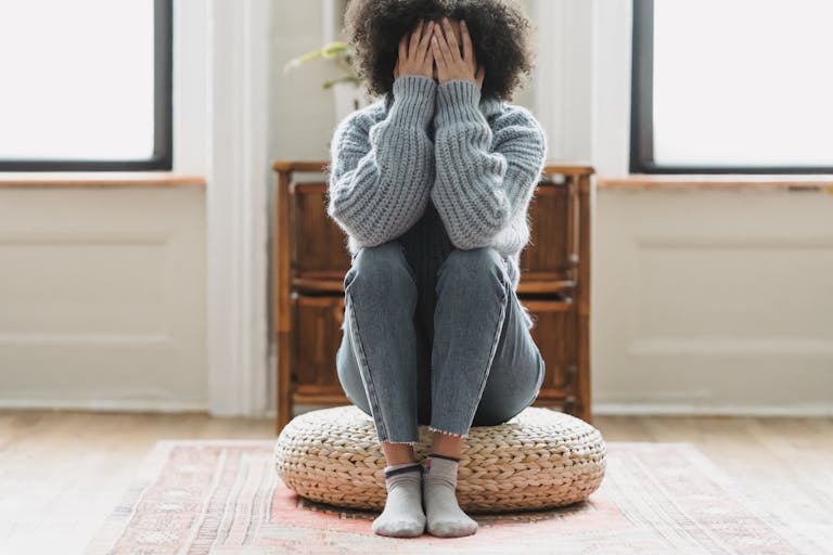 Full body of anonymous depressed female in casual clothes sitting on rattan stool in light room with windows at home