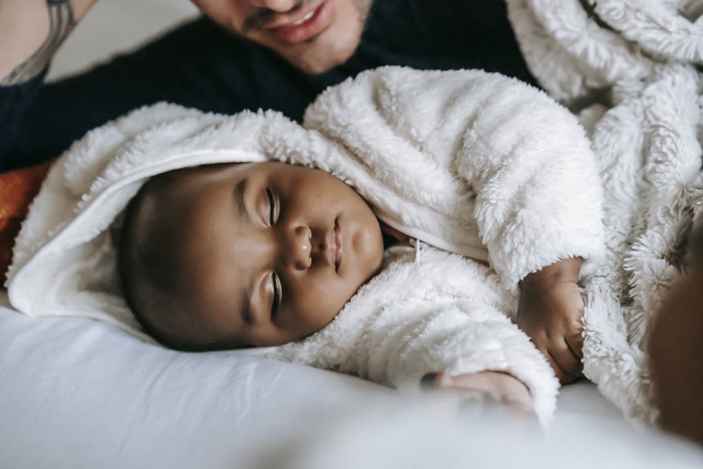 Unrecognizable ethnic father lying on bed with African American newborn baby in white pajama sleeping in bedroom during bed time at home