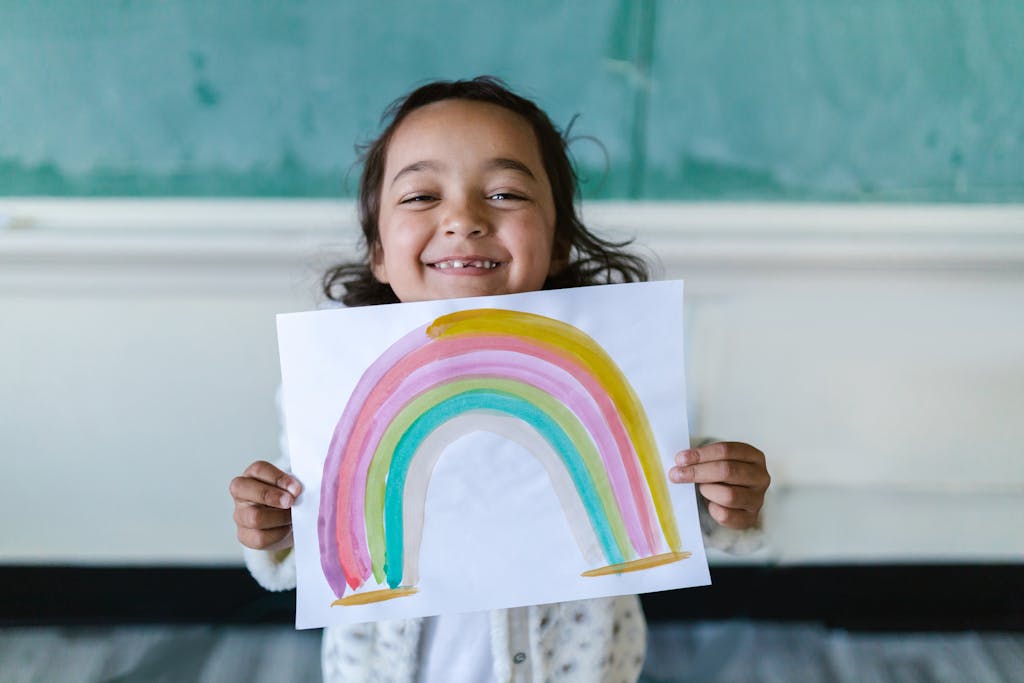 A cheerful child proudly displays a watercolor rainbow painting in a classroom setting.